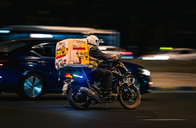 A delivery motorbike speeds through the streets of Kuwait City at night, showcasing fast-paced urban life.